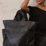 A large black pebbled leather tote bag with top handles and side pockets, shown next to a woman wearing a black off-the-shoulder top against a textured light gray wall.