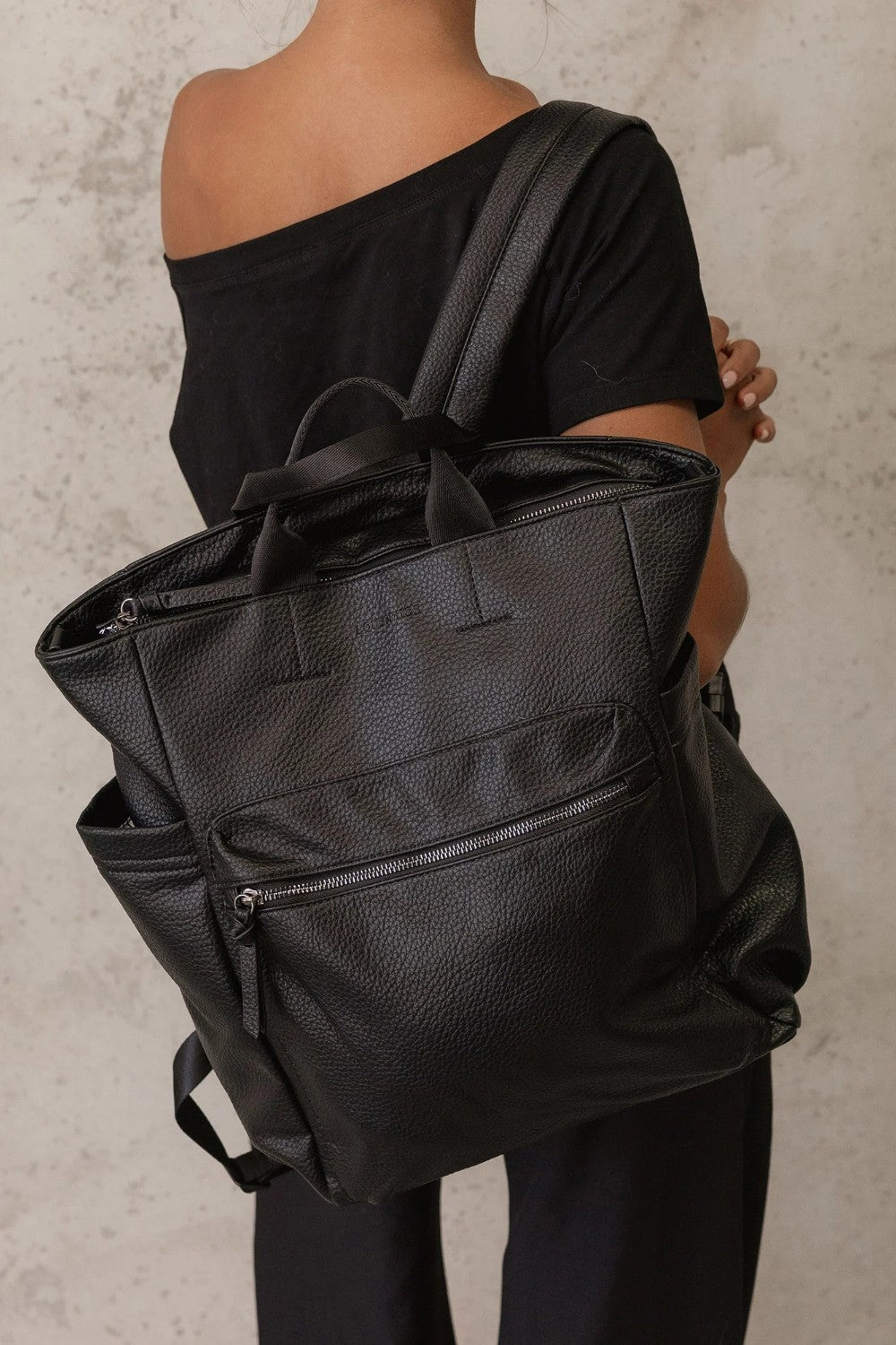 A black pebbled faux-leather backpack with top handles and side pockets, shown from the back on a model wearing a black off-the-shoulder top against a textured light gray wall.