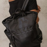 A black pebbled faux-leather backpack with top handles and side pockets, shown from the back on a model wearing a black off-the-shoulder top against a textured light gray wall.