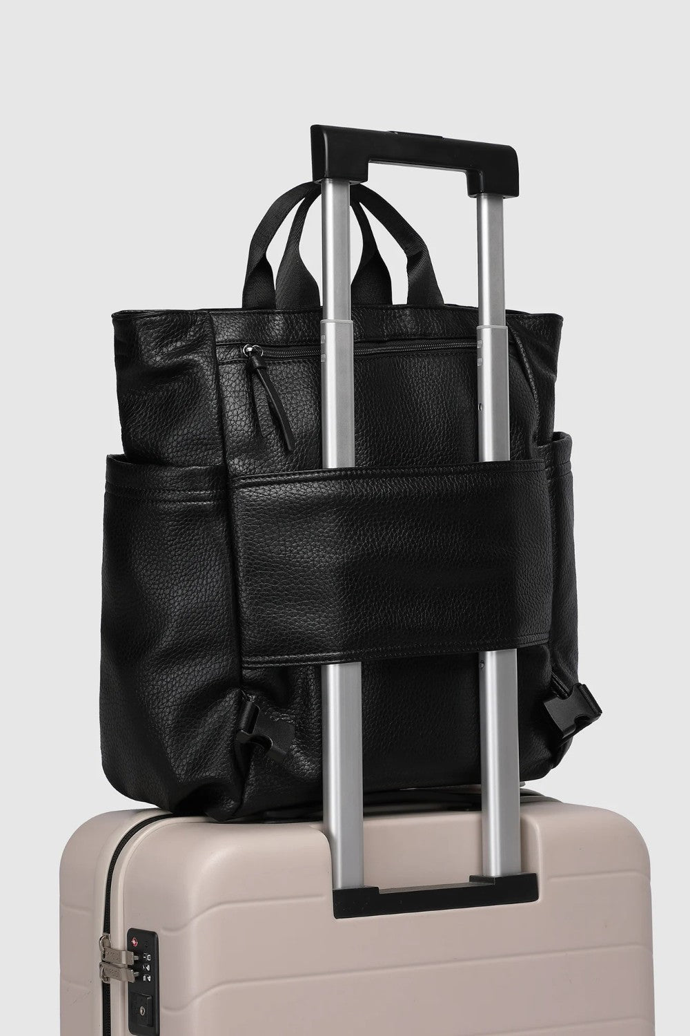 A black pebbled faux-leather backpack with an extended telescoping handle, shown resting on top of a light beige hard-shell suitcase against a light gray background.