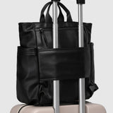 A black pebbled faux-leather backpack with an extended telescoping handle, shown resting on top of a light beige hard-shell suitcase against a light gray background.