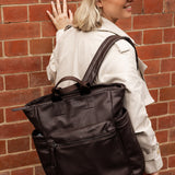 Person wearing a brown cap and carrying a brown backpack against a brick wall.
