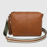 A tan leather crossbody bag with a green and white striped woven strap, featuring a brown leather tassel charm on the side, set against a light gray background.