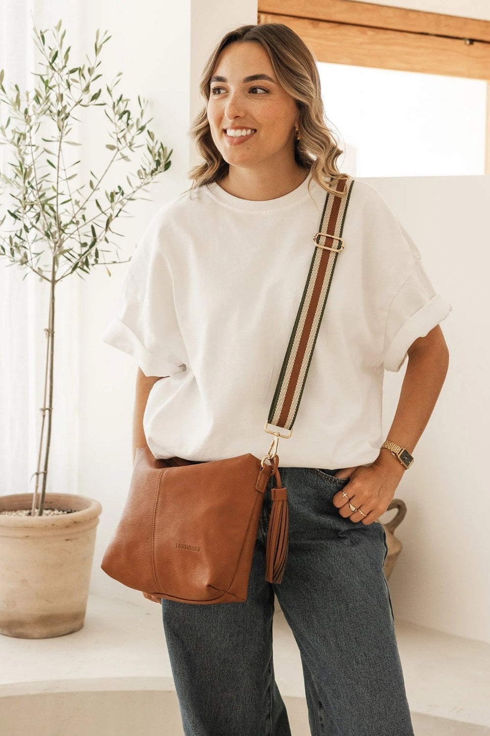 A woman wearing a white t-shirt and dark jeans models a brown leather crossbody bag with a striped strap in a bright, modern room with a potted plant.