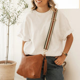 A woman wearing a white t-shirt and dark jeans models a brown leather crossbody bag with a striped strap in a bright, modern room with a potted plant.