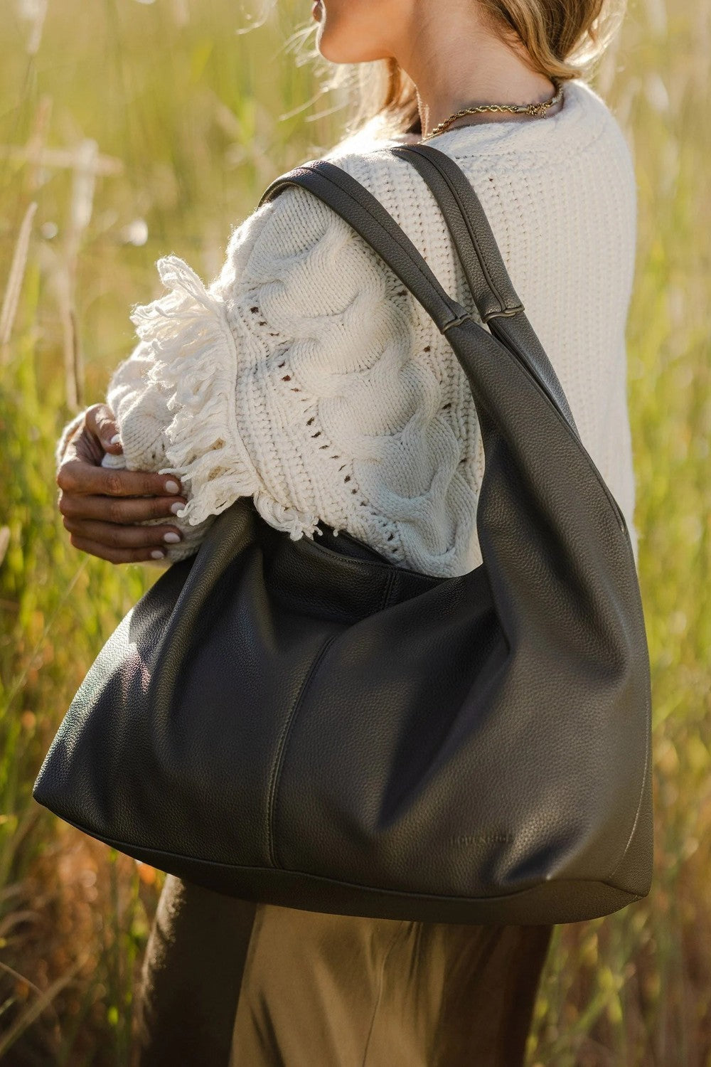 A woman outdoors in a grassy field carrying a large olive green leather hobo shoulder bag over her shoulder. She is wearing a white textured knit top and olive pants.