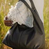 A woman outdoors in a grassy field carrying a large olive green leather hobo shoulder bag over her shoulder. She is wearing a white textured knit top and olive pants.