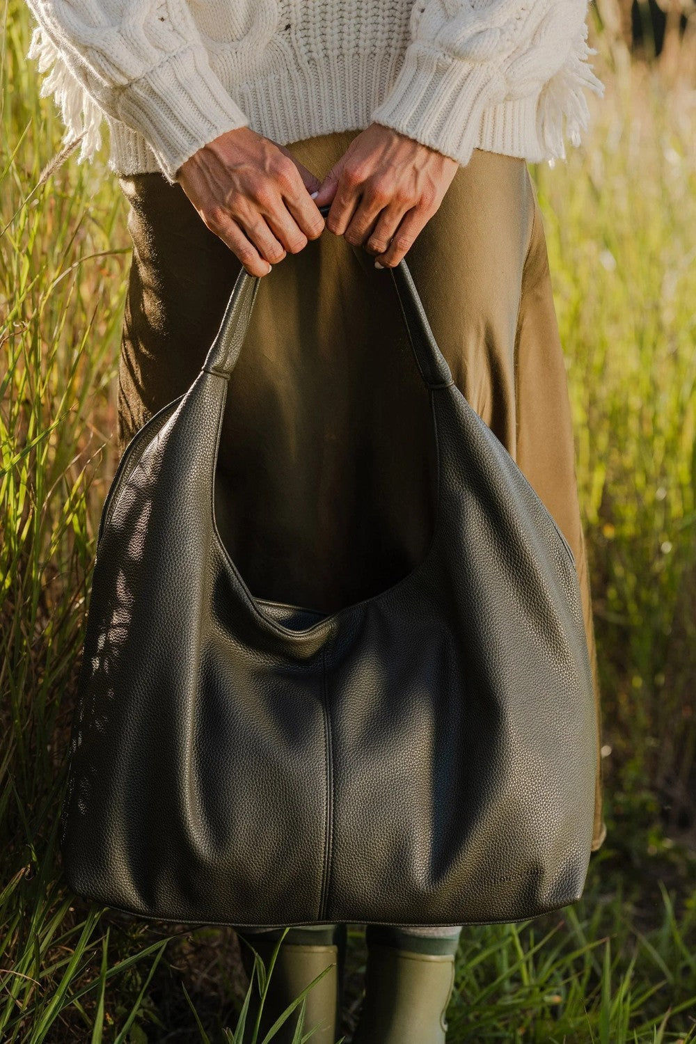 A person outdoors in a grassy field holding a large slouchy shoulder bag made of dark olive green pebbled leather. The person is wearing a cream cable-knit sweater, brown trousers, and green rain boots.