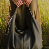 A person outdoors in a grassy field holding a large slouchy shoulder bag made of dark olive green pebbled leather. The person is wearing a cream cable-knit sweater, brown trousers, and green rain boots.