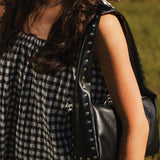 A close-up lifestyle shot of a woman wearing a black and white gingham dress and carrying a black leather shoulder bag adorned with silver studs.