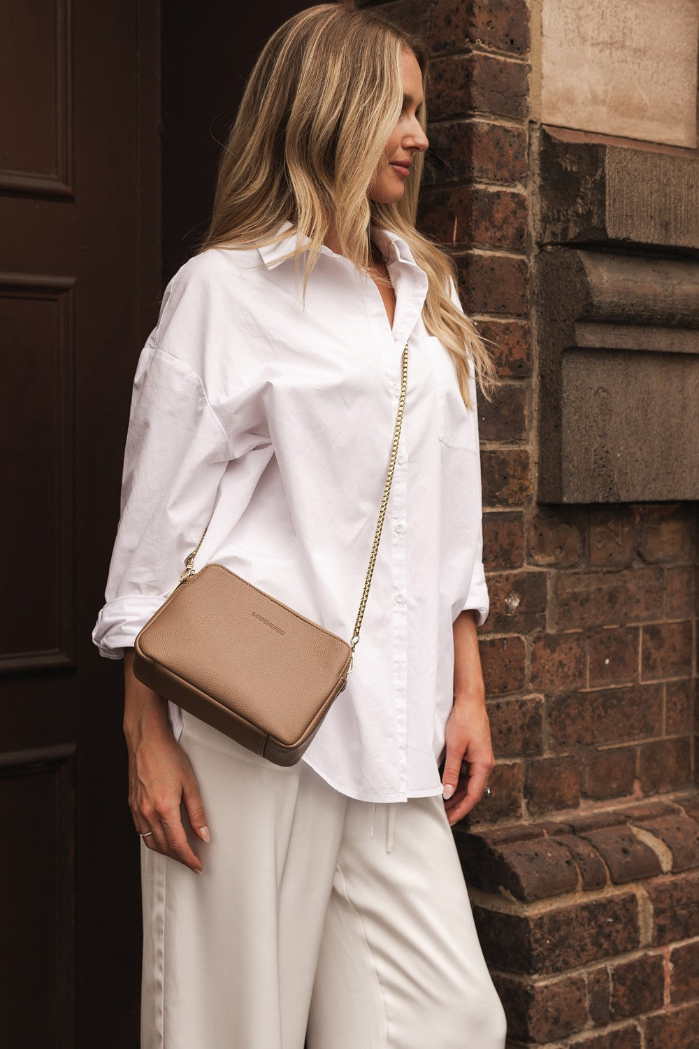 Woman in a white shirt and beige pants holding a beige handbag against a brick wall.