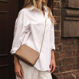 Woman in a white shirt and beige pants holding a beige handbag against a brick wall.