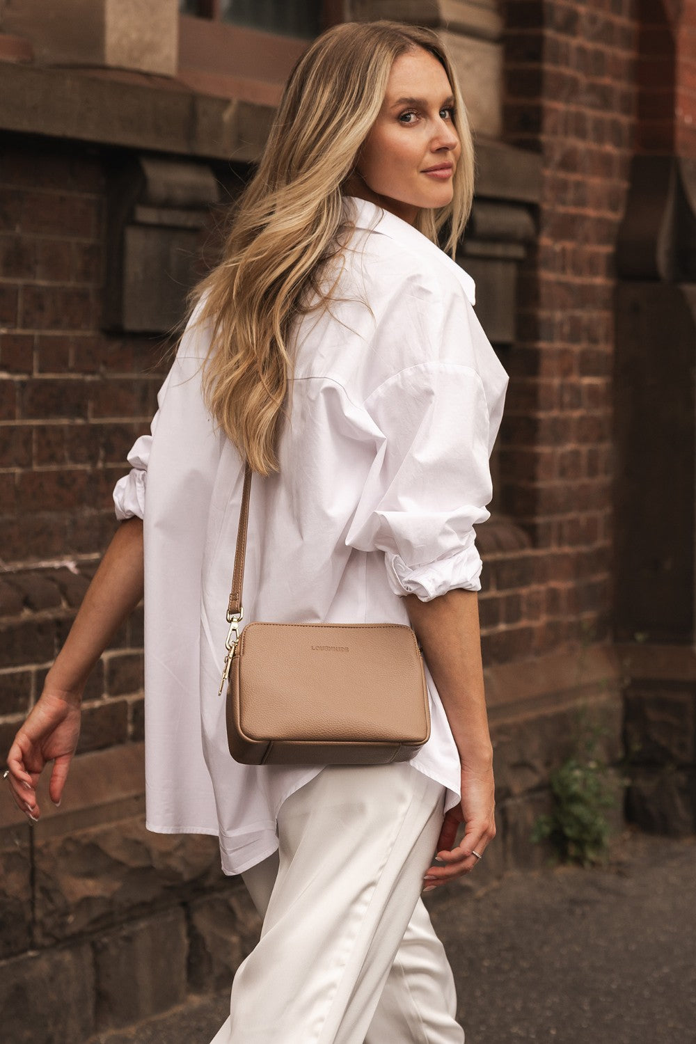 Woman in a white outfit with a beige handbag standing against a brick wall.