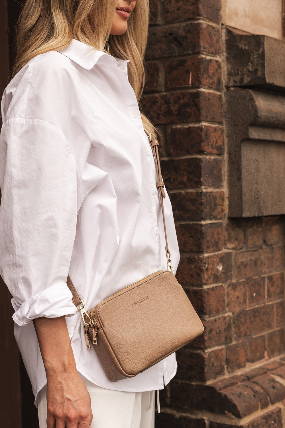 Person wearing a white shirt with a beige handbag against a brick wall.