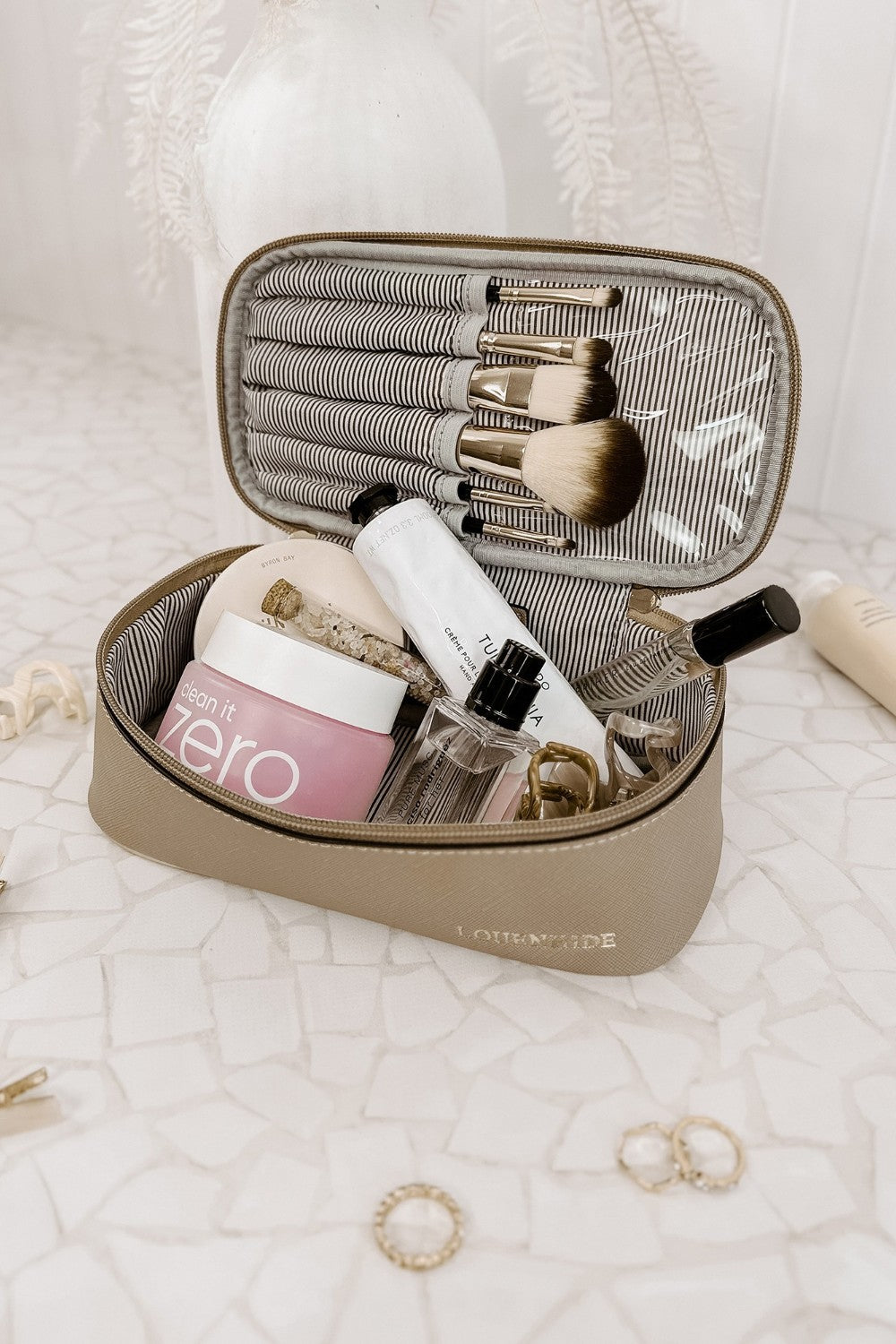 Makeup set with brushes, bottles, and jars in a beige travel case on a textured white surface.