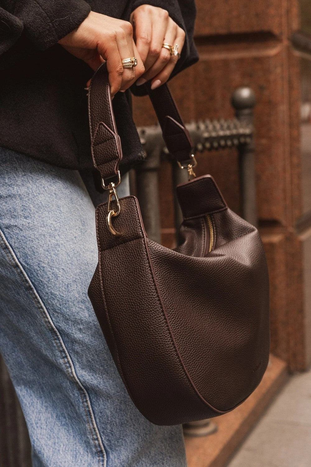 A person wearing a brown top and light blue jeans carries a dark brown pebbled leather shoulder bag with gold-tone hardware in an outdoor urban setting.