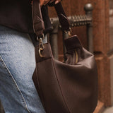 A person wearing a brown top and light blue jeans carries a dark brown pebbled leather shoulder bag with gold-tone hardware in an outdoor urban setting.
