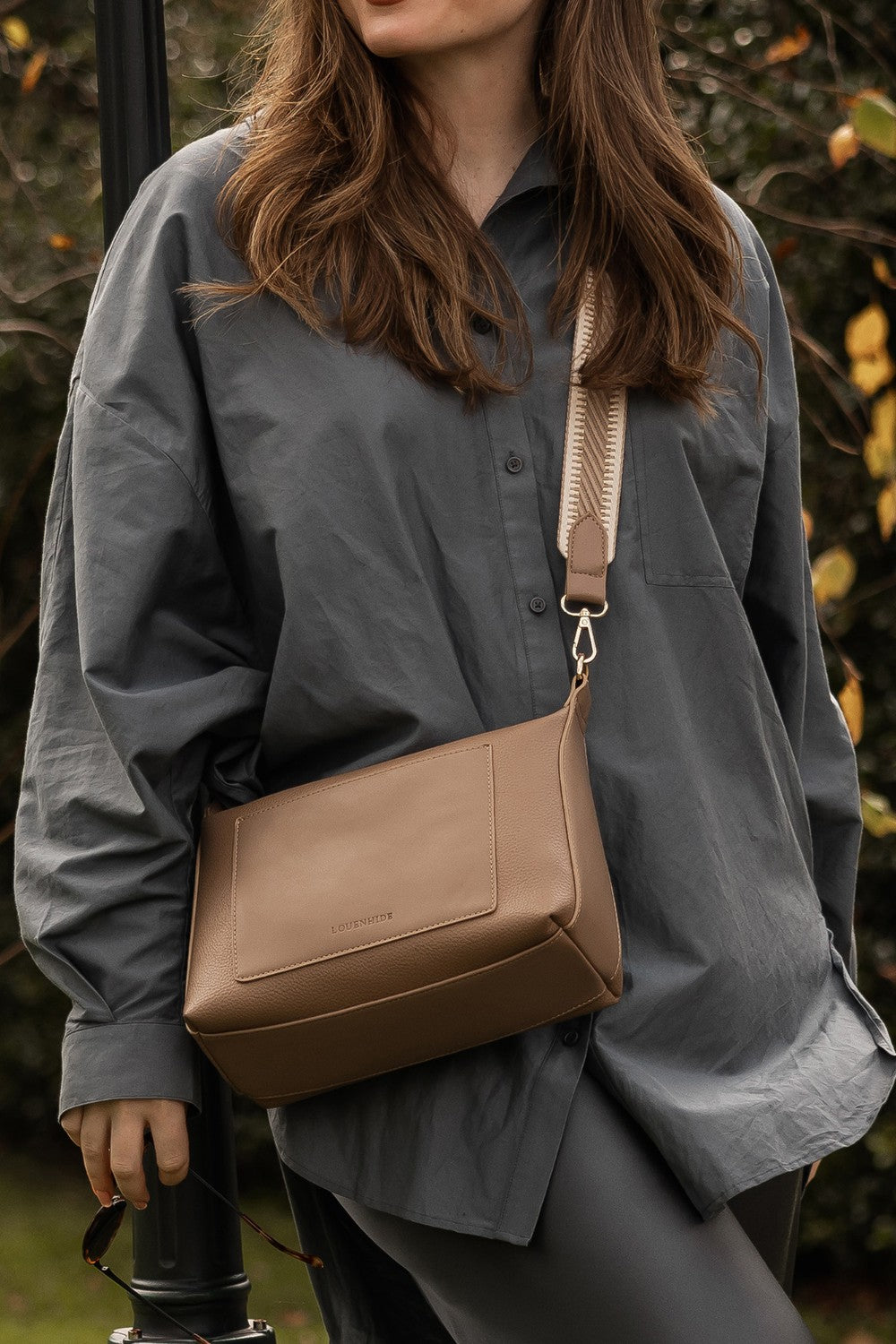 Person wearing a gray shirt with a brown pleather bag over their shoulder against a natural background.