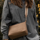 Person wearing a gray shirt with a brown pleather bag over their shoulder against a natural background.