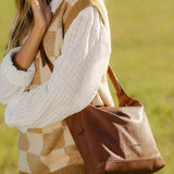 A woman outdoors in a grassy field wearing a cream and tan argyle sweater vest over a white shirt, carrying a large brown leather shoulder bag.