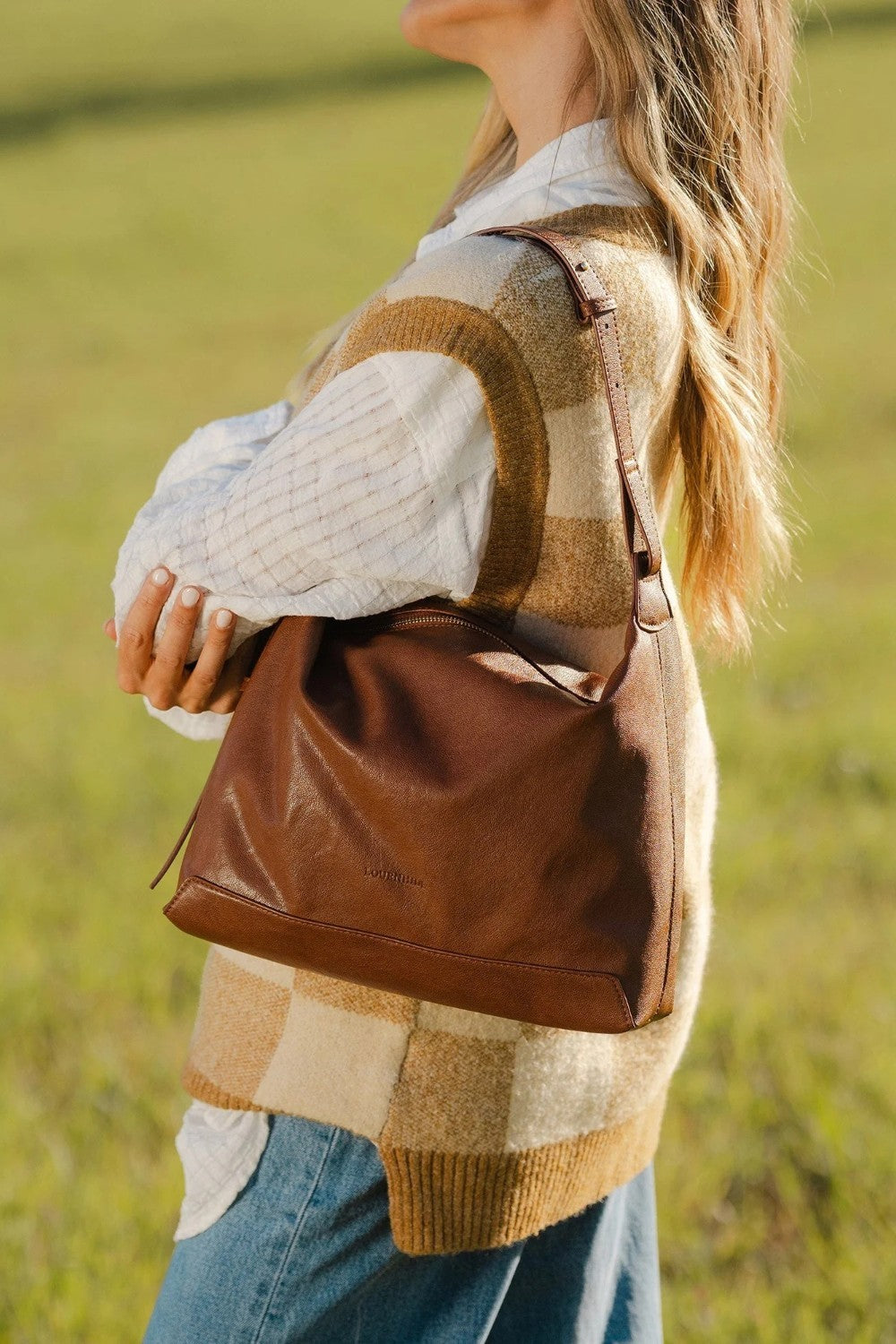 Person wearing a plaid sweater and holding a brown leather bag in a grassy field