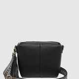 A black pebbled leather crossbody bag with a zip-top closure, a tassel detail, and a wide woven strap with silver-tone hardware, shown against a light gray background.