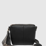 A black pebbled leather crossbody bag with a zippered top and a wide woven strap plus an additional black pleather strap, shown against a light gray background.