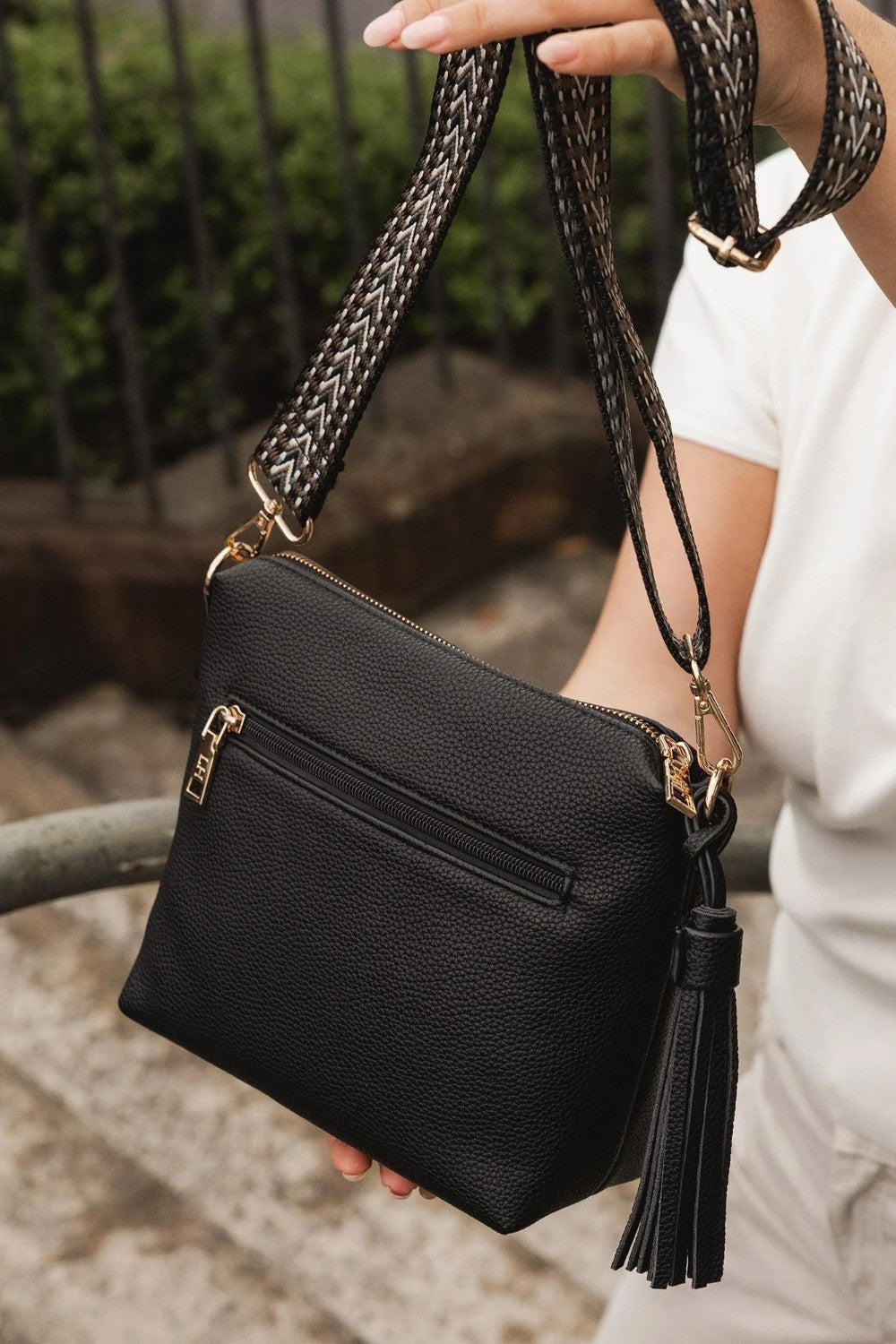 A black pebbled faux-leather crossbody bag with gold-tone hardware, held by a person wearing a white top. The bag features a shoulder strap, a external zip pocket, and a black tassel charm.