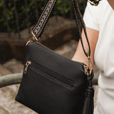 A black pebbled faux-leather crossbody bag with gold-tone hardware, held by a person wearing a white top. The bag features a shoulder strap, a external zip pocket, and a black tassel charm.