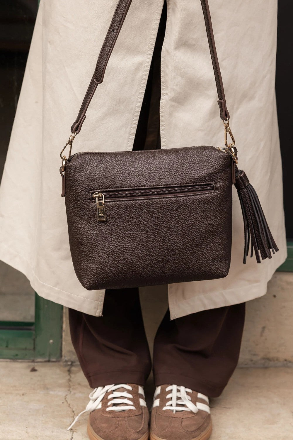 A dark brown pebbled leather crossbody bag with an external zip pocket and a tassel zipper pull, shown being worn by a person in a beige trench coat and brown pants.