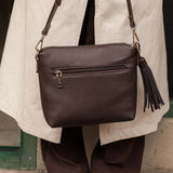 A dark brown pebbled leather crossbody bag with an external zip pocket and a tassel zipper pull, shown being worn by a person in a beige trench coat and brown pants.