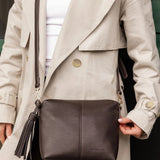 A dark brown pebbled leather crossbody bag with a tassel charm, worn by a model in a beige trench coat over a white top.