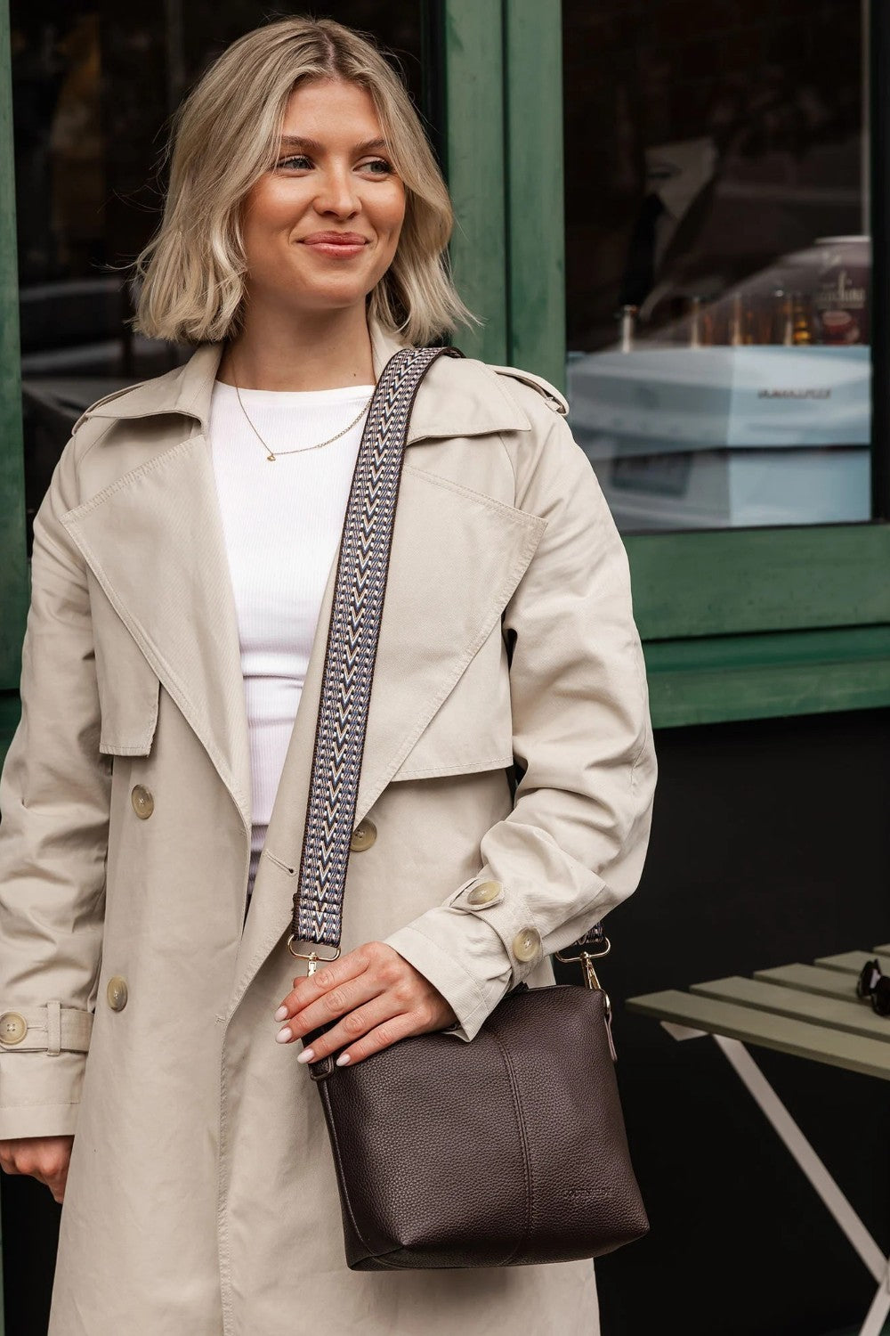 A woman wearing a beige trench coat over a white top, holding a dark brown leather shoulder bag with a patterned strap.