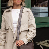 A woman wearing a beige trench coat over a white top, holding a dark brown leather shoulder bag with a patterned strap.