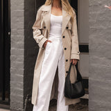 Woman in a beige trench coat and white outfit standing against a building.