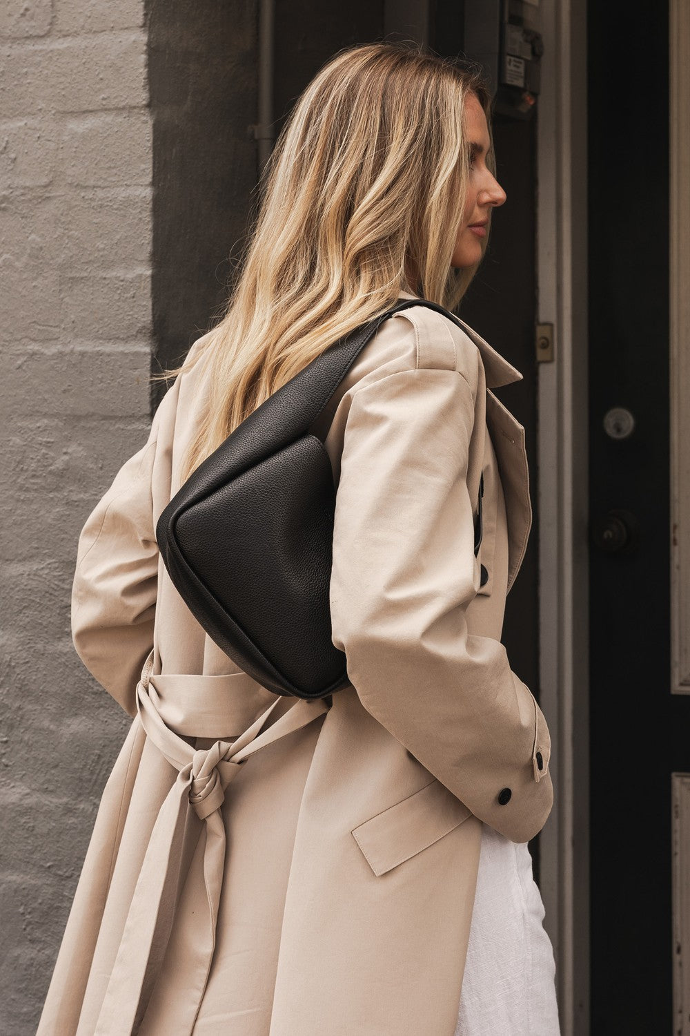 Woman in a beige trench coat with a black bag walking past a door.