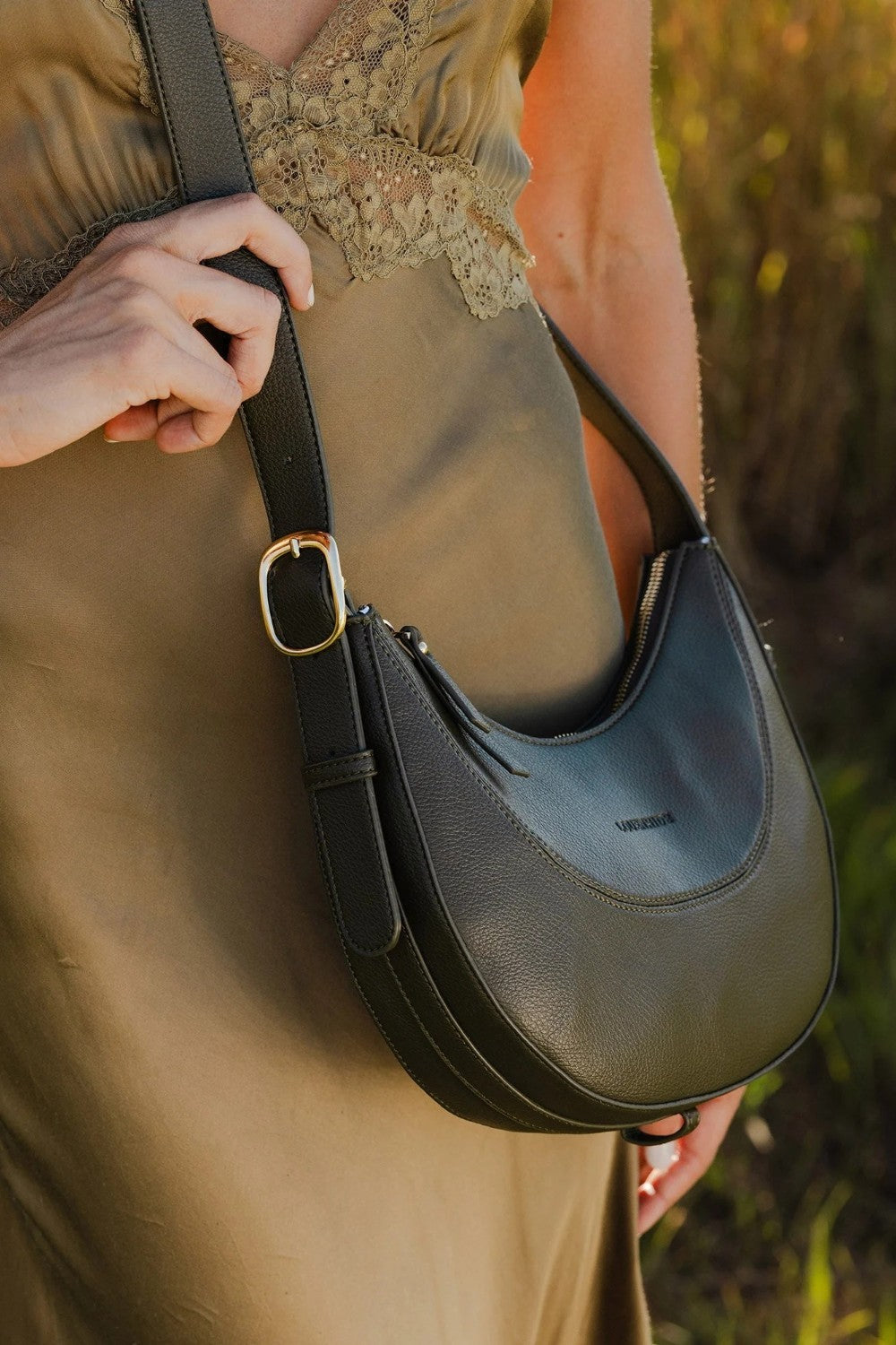 A person wearing a green dress carries a dark olive green crescent-shaped shoulder bag with a gold buckle, set against a blurred outdoor background.