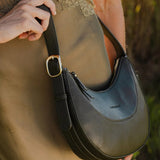 A person wearing a green dress carries a dark olive green crescent-shaped shoulder bag with a gold buckle, set against a blurred outdoor background.