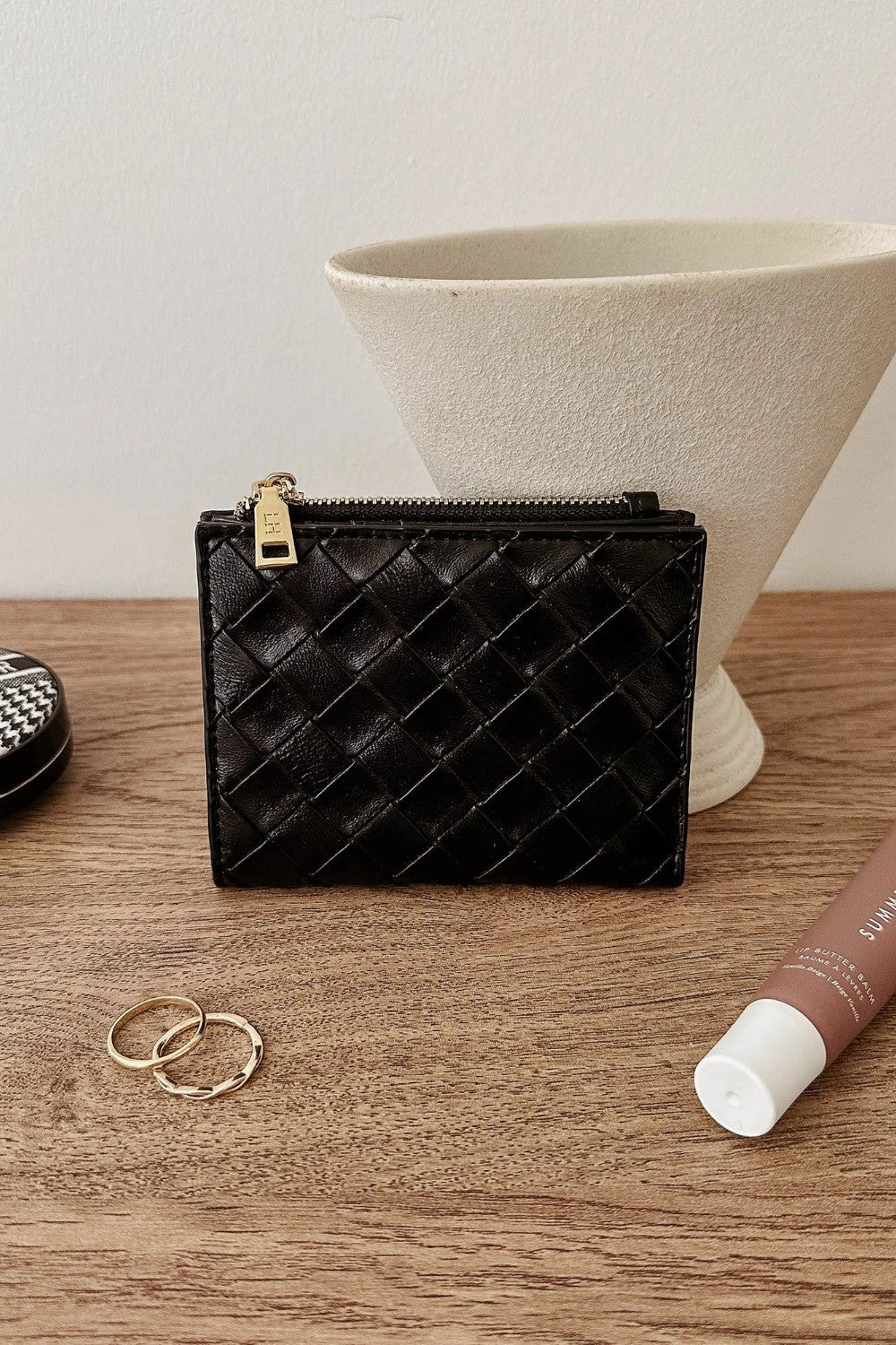 A black woven leather zip-around card case with gold-tone hardware, displayed on a wooden tabletop with decorative props including rings, a tube, and a ceramic vase.