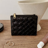 A black woven leather zip-around card case with gold-tone hardware, displayed on a wooden tabletop with decorative props including rings, a tube, and a ceramic vase.