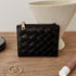A black woven leather zip-around card case with gold-tone hardware, displayed on a wooden tabletop with decorative props including rings, a tube, and a ceramic vase.