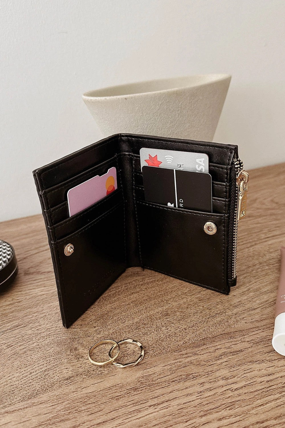 A black leather bifold wallet shown open on a wooden tabletop, with card slots, a zipper pocket, and small gold-tone hardware visible. A cream vase and rings are in the background and surroundings.