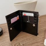A black leather bifold wallet shown open on a wooden tabletop, with card slots, a zipper pocket, and small gold-tone hardware visible. A cream vase and rings are in the background and surroundings.