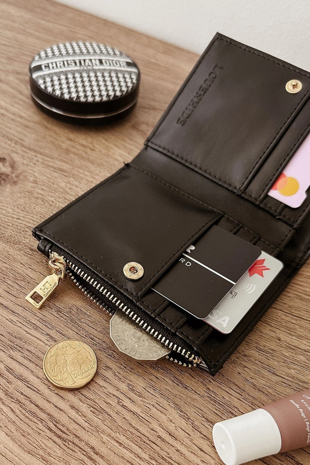 A black leather bifold wallet shown open on a wooden tabletop, with cards, coins, and a small round compact nearby. The wallet has gold-tone hardware and a visible interior embossed LOUENHIDE logo.