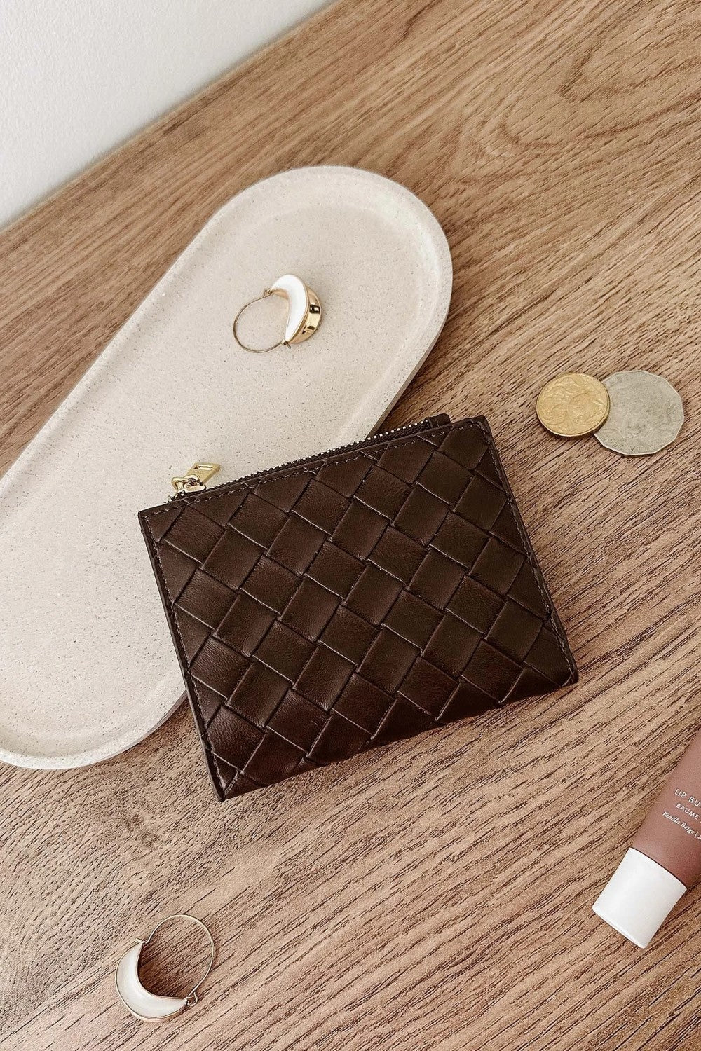 A dark brown woven leather zip pouch wallet, styled on a light wood tabletop next to a cream-colored jewelry tray, coins, and rings.