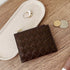 A dark brown woven leather zip pouch wallet, styled on a light wood tabletop next to a cream-colored jewelry tray, coins, and rings.