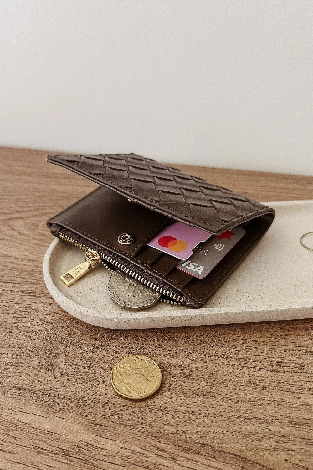 A dark brown woven leather wallet with a zippered compartment, shown open on a light beige tray over a wooden surface. Credit cards and coins are partially visible inside the wallet.