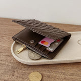 A dark brown woven leather wallet with a zippered compartment, shown open on a light beige tray over a wooden surface. Credit cards and coins are partially visible inside the wallet.