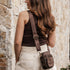 Woman with a brown tank top and beige skirt holding a brown crossbody bag against a stone wall.