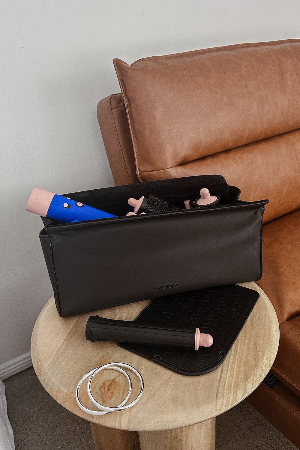 Black travel case with makeup items on a wooden side table next to a brown leather sofa.